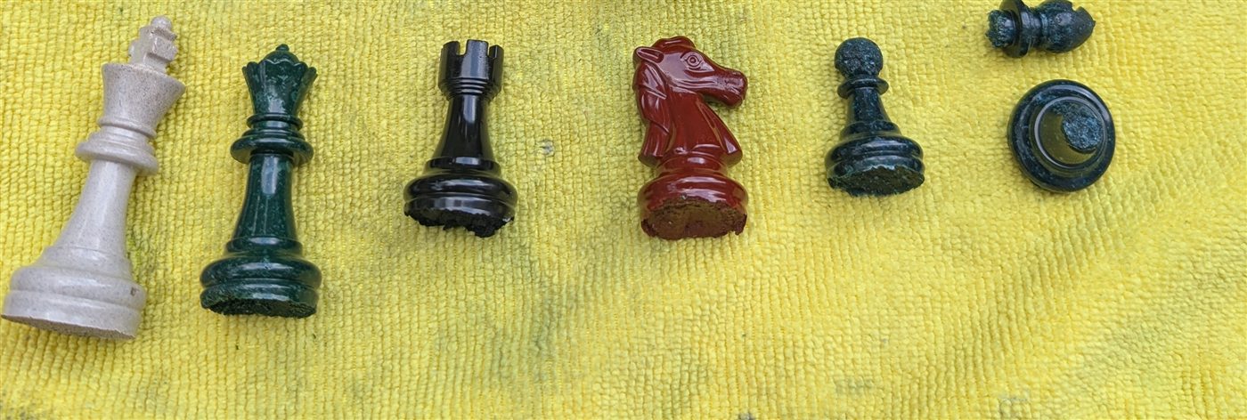Hand-cast chess pieces made from cremains blend: a bone-white king, deep green queen, glossy black rook, oxblood red knight and green pawn.